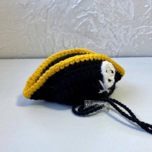 May include: A black crocheted pirate hat with a yellow trim and a white skull and crossbones design. The hat has a black string attached to it.