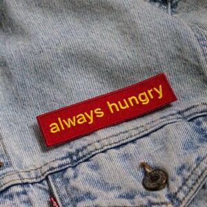 Puede incluir: Una chaqueta vaquera roja con un parche rojo que dice "always hungry" en bordado amarillo.