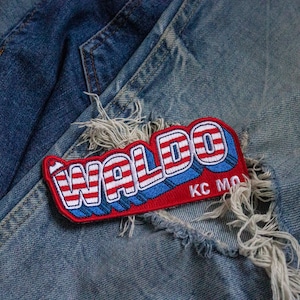 May include: A red, white, and blue embroidered patch with the word "WALDO" in a retro font. The patch has "KC MO" written in smaller letters below the word "WALDO". The patch is attached to a pair of blue denim jeans.