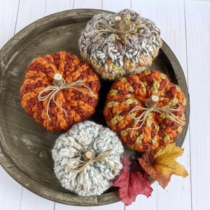 May include: Four crocheted pumpkins in various fall colors, including orange, brown, and white, are displayed on a wooden tray. The pumpkins are decorated with twine and have a rustic, handmade look.