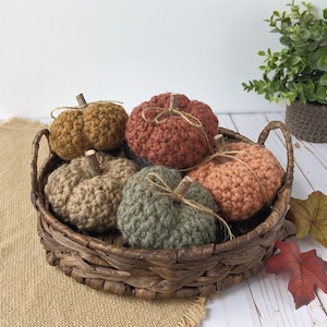 May include: Five crocheted pumpkins in a brown wicker basket. The pumpkins are in shades of orange, brown, and green.