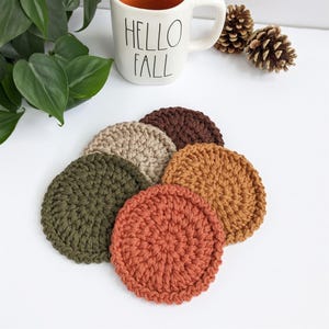 May include: A collection of six handmade, circular crochet coasters in various fall colors: olive green, tan, brown, rust, and orange. A white mug with "HELLO FALL" printed on it and two pinecones are in the background.
