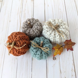 May include: Four crocheted pumpkins in different colors, including brown, orange, blue, and white, with wooden stems. The pumpkins are arranged on a white wooden surface with fall leaves.