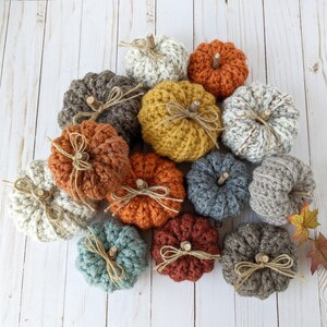 May include: A collection of 13 crocheted pumpkins in various colors, including beige, brown, orange, yellow, and blue. The pumpkins are arranged in a circle on a white wooden surface.
