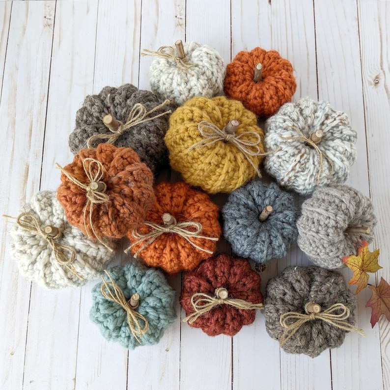 May include: A collection of 13 crocheted pumpkins in various colors, including beige, brown, orange, yellow, and blue. The pumpkins are arranged in a circle on a white wooden surface.