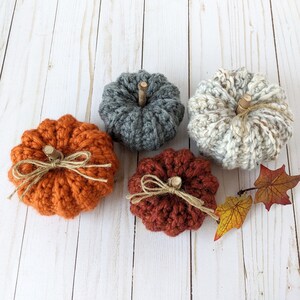 May include: Four crocheted pumpkins in different colors and styles. The pumpkins are on a white wooden background.