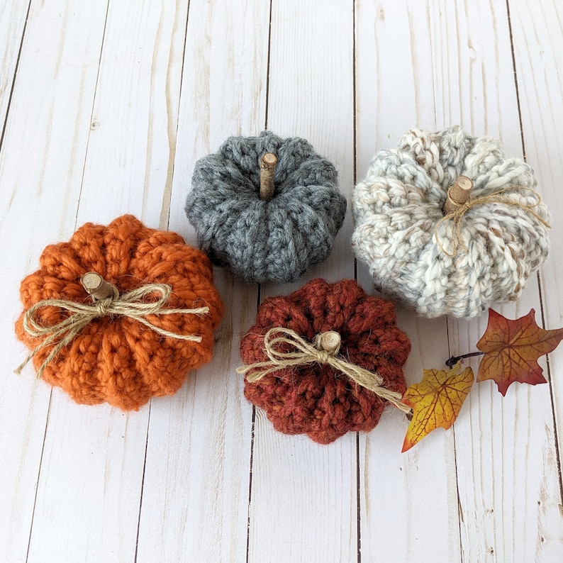 May include: Four crocheted pumpkins in different colors and styles. The pumpkins are on a white wooden background.
