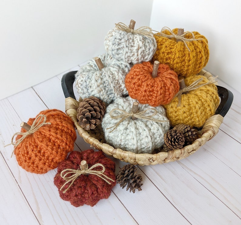 May include: A collection of crocheted pumpkins in various colors, including orange, white, and yellow, are displayed in a woven basket. The pumpkins are decorated with twine and have a rustic, autumnal feel.