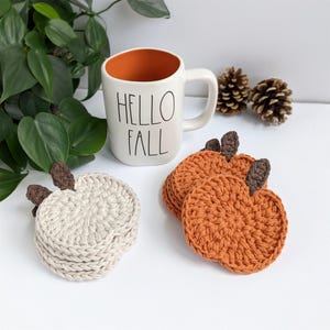 May include: A white mug with "HELLO FALL" printed on it, next to a stack of cream-colored and orange crocheted pumpkin coasters. The coasters have brown stem details. Two pinecones and green leaves are in the background.