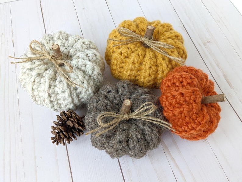 May include: Four crocheted pumpkins in various colors, including white, yellow, orange, and brown. Each pumpkin has a small wooden stem and a twine bow.