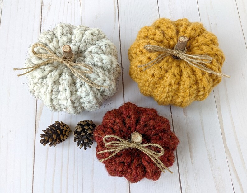 May include: Three crocheted pumpkins in white, yellow, and red. The pumpkins are decorated with twine bows and wooden stems.