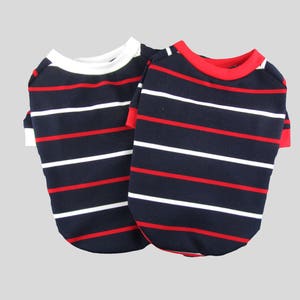 Puede incluir: Dos camisas a rayas para perros, una con cuello blanco y otra con cuello rojo. Ambas camisas tienen rayas azul marino y rojas.