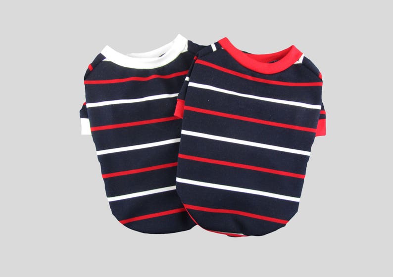 Puede incluir: Dos camisas a rayas para perros, una con cuello blanco y otra con cuello rojo. Ambas camisas tienen rayas azul marino y rojas.