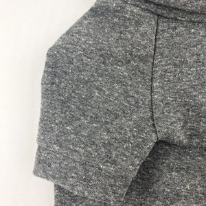 May include: A close-up of the gray heather fabric of a short-sleeved shirt. The fabric has a textured, slightly speckled appearance.