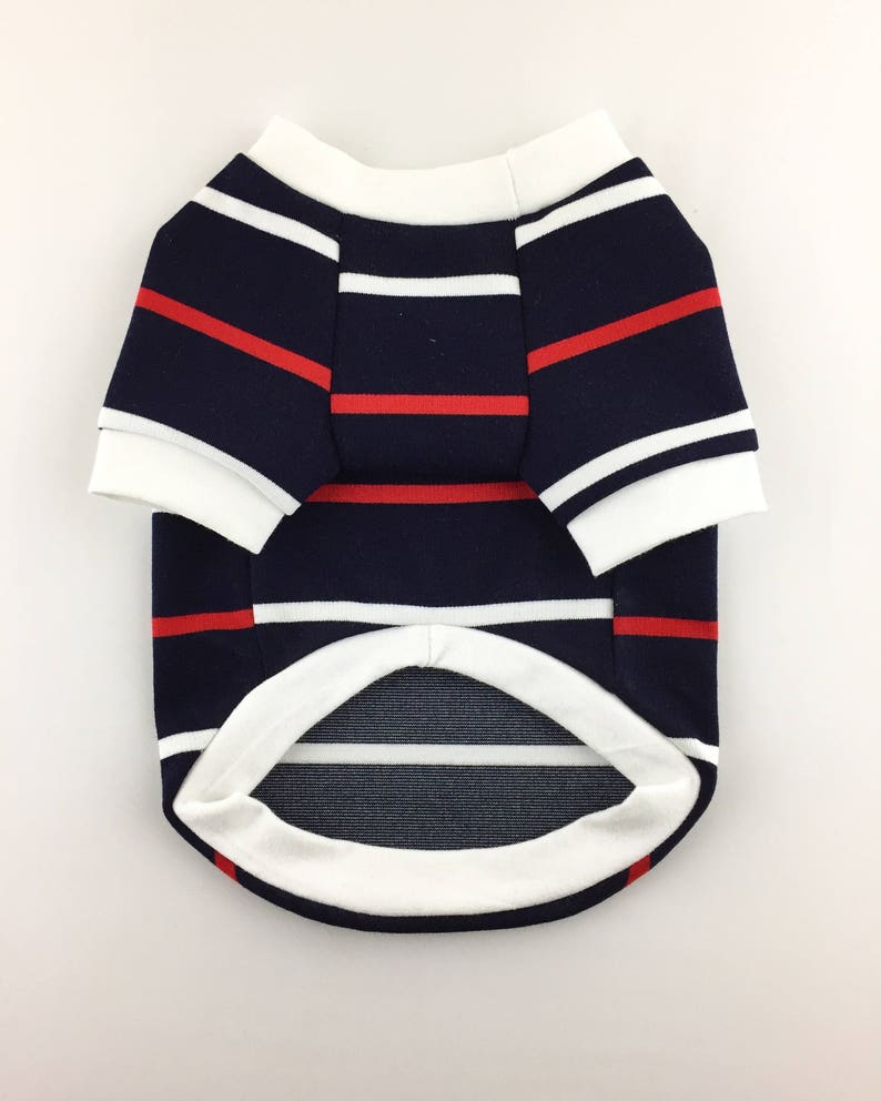 Puede incluir: Una camiseta para perro de rayas azul marino y blancas con rayas rojas. La camiseta tiene un cuello y pu&ntilde;os blancos.