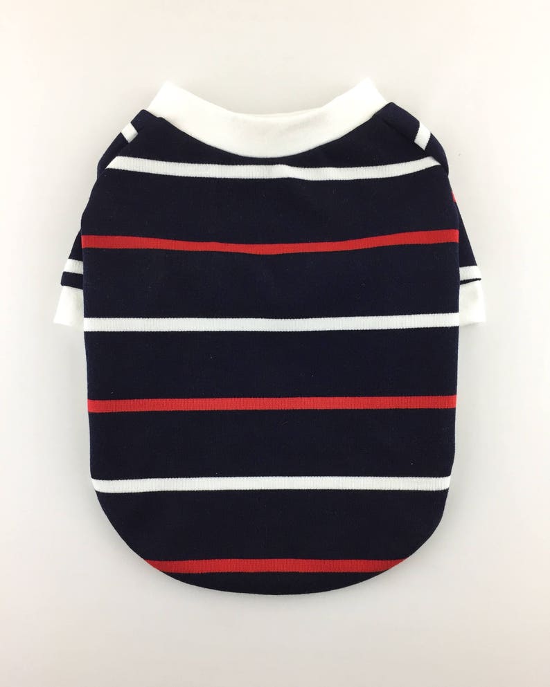 Puede incluir: Una camiseta para perro a rayas azul marino y blancas con rayas rojas. La camiseta tiene un cuello blanco.