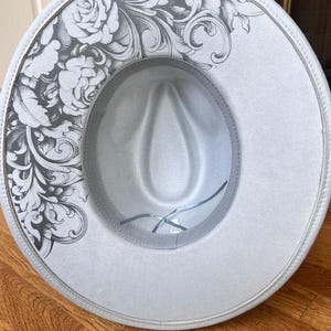 May include: A light gray fedora hat with a decorative floral design on the brim. The hat has a structured crown and a contrasting gray interior. The brim is approximately 3 inches wide.