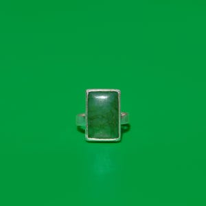 Op de afbeelding: Een rechthoekige groene edelsteenring met een zilveren band. De edelsteen heeft een diepgroene kleur en is gezet in een zilveren bezel. De ring is gecentreerd op een groene achtergrond.