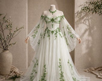 May include: A white wedding dress with green leaf embroidery. The off-the-shoulder gown has long, flowing sleeves and a full skirt. A matching green necklace is on the mannequin. The dress is set against a neutral backdrop.