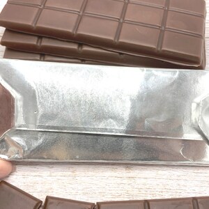May include: A silver foil-wrapped chocolate bar with a folded edge. The chocolate bar is sitting on a white wooden surface.