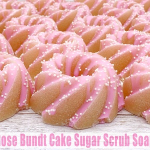 May include: A close-up of several pink and white sugar scrub soaps shaped like bundt cakes. The soaps are arranged in rows on a white wooden surface. The text "Rose Bundt Cake Sugar Scrub Soap" is printed below the soaps.