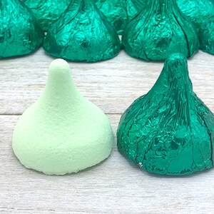 May include: Two green chocolate kisses, one wrapped in foil and one unwrapped, on a light wood surface.