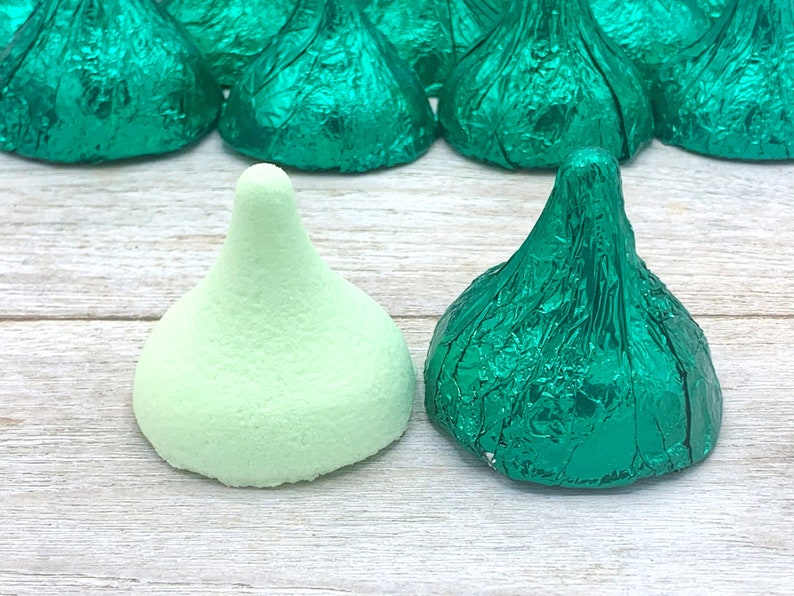 May include: Two green chocolate kisses, one wrapped in foil and one unwrapped, on a light wood surface.