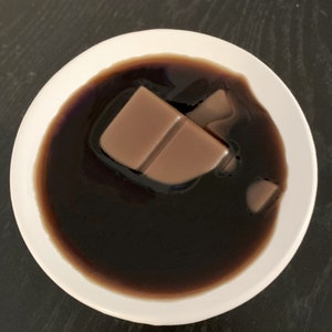 May include: A white bowl filled with dark brown liquid and a broken piece of chocolate bar floating on top.