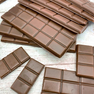 May include: A stack of milk chocolate bars on a white wooden surface. The bars are arranged in a staggered pattern, with the top bar being the largest and the bottom bar being the smallest. The bars are all the same size and shape, and they are all made of milk chocolate.
