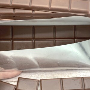 May include: A chocolate bar wrapped in silver foil. The bar is divided into squares and is partially unwrapped, revealing the dark brown chocolate inside.