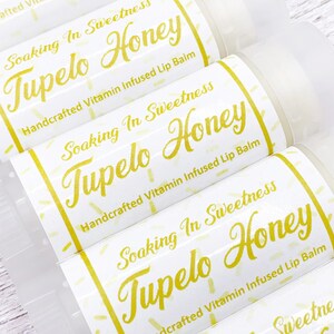 May include: Close-up of four lip balm tubes with white labels and yellow text. The labels read "Soaking In Sweetness Tupelo Honey Handcrafted Vitamin Infused Lip Balm".