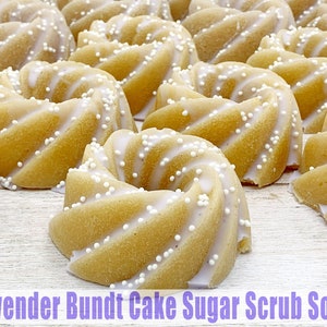 May include: A close-up of several small, white and yellow bundt cake shaped sugar scrub soaps. The soaps are topped with white sprinkles and have a lavender scent.