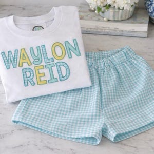 May include: A white t-shirt and matching shorts set. The shirt features the name "WAYLON REID" in blue, yellow, and checkered letters. The shorts are light blue and white checkered.