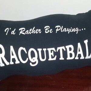 Puede incluir: Una almohada negra con texto blanco que dice "I'd Rather Be Playing... Racquetball".