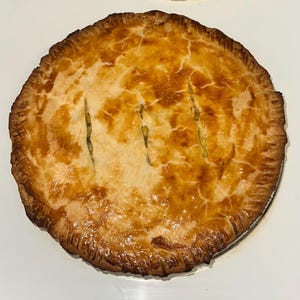 Può includere: Una torta rotonda dorata con una crosta friabile. La torta ha tre fessure distanziate sulla parte superiore, rivelando parte del ripieno. La crosta è increspata attorno ai bordi e la torta è su una superficie bianca.