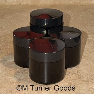 May include: A group of five small, black cosmetic jars with matching black lids. The jars are arranged in a circular pattern, with one jar stacked on top. The text "©M Turner Goods" is visible at the bottom.