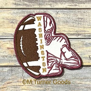 May include: A maroon and brown football-shaped patch featuring a football player and the word "WASHINGTON" in gold lettering. The patch is on a wooden background.