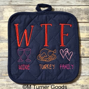 May include: Navy blue quilted pot holder with the embroidered letters "WTF" in red. Below the letters are embroidered images of wine glasses, a turkey, and hearts, with the words "WINE", "TURKEY", and "FAMILY".
