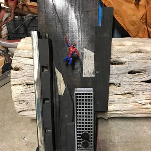 May include: A mixed media artwork featuring a Spider-Man action figure clinging to a dark wooden structure. The composition includes various geometric shapes, metal elements, and a textured wooden beam, creating a unique assemblage.