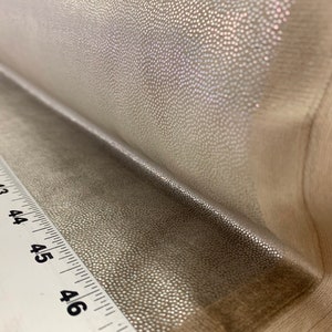 May include: Close-up of a silver fabric with a textured, pebbled surface. The fabric is folded over a white ruler with black markings.