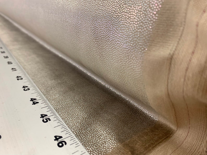 May include: Close-up of a silver fabric with a textured, pebbled surface. The fabric is folded over a white ruler with black markings.