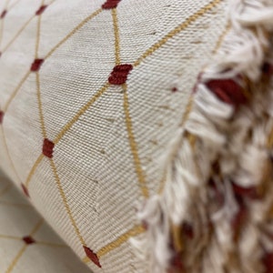 May include: Close-up of a white fabric with a geometric pattern of gold lines and red squares. The fabric has a frayed edge.