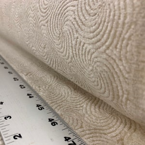 May include: Close-up of a white fabric with a swirling pattern. The fabric is rolled up and a ruler is visible in the foreground.