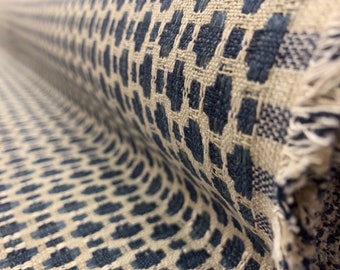 May include: A roll of woven fabric featuring a repeating geometric pattern in dark blue and off-white. The textile has a textured surface with visible interlaced threads and a frayed edge showing the raw construction of the material.