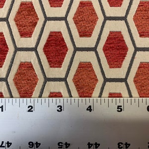 May include: Close-up of a fabric with a geometric pattern of red hexagons on a cream background. The fabric is textured and has a subtle sheen. A ruler is placed at the bottom of the image, showing the fabric is 6 inches wide.