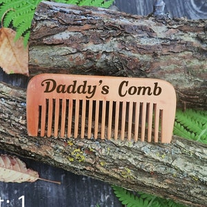 Könnte beinhalten: Ein Holzkamm mit der Gravur "Daddy's Comb". Der Kamm liegt auf einem Stück Baumrinde.