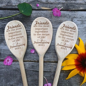 May include: Three wooden spoons with engraved text: "Friends are the most important ingredient in the recipe of life!" and "Friendsgiving 2021." The spoons are arranged on a rustic wooden surface, with flowers and leaves.