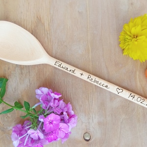 May include: A wooden spoon engraved with "Edward + Rebecca" and a heart symbol, followed by the date "14.02.2021". The spoon is surrounded by pink and yellow flowers on a wooden surface.