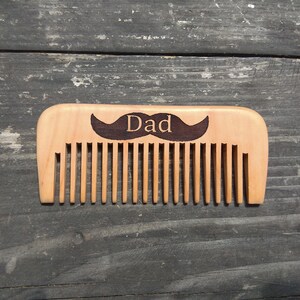 Puede incluir: Peine de madera con un diseño de bigote y la palabra "Papá" grabada.