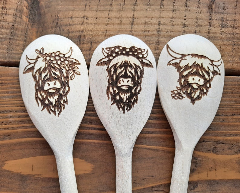 Puede incluir: Tres cucharas de madera con un dise&ntilde;o de vaca Highland quemado en la madera. Las cucharas son blancas con un dise&ntilde;o marr&oacute;n.
